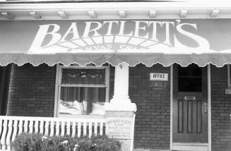 Shows a brick house with a large awning that reads "Bartlett's". A sign on the verandah says: W ...