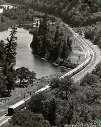 Shows the Agawa Canyon tour train, on a curve, with forest to one side and the river to the oth ...