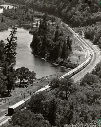 Shows the Agawa Canyon tour train, on a curve, with forest to one side and the river to the oth…