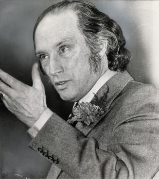 A close-up of a man with longish hair and side whiskers, wearing a carnation, speaking intently ...