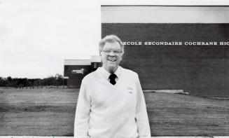 Smiling middle-aged white man wearing a tie and glasses, standing in front of a Catholic high s…