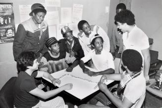 Nine smiling young people sitting around a table playing dominoes.