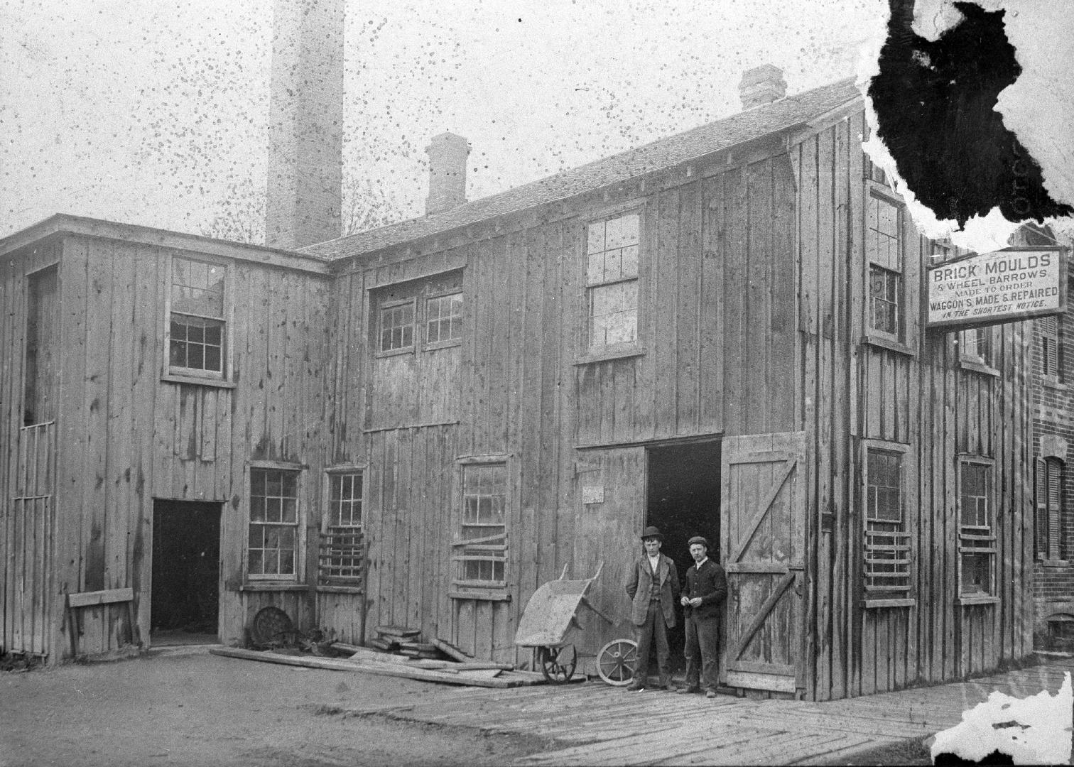 Image shows a few people standing beside open doors of brickmakers' supplies building.