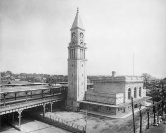 Image shows a rail station building with a clock tower, train platform and tracks.