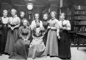 Nine women in long dresses, with long hair tied up.