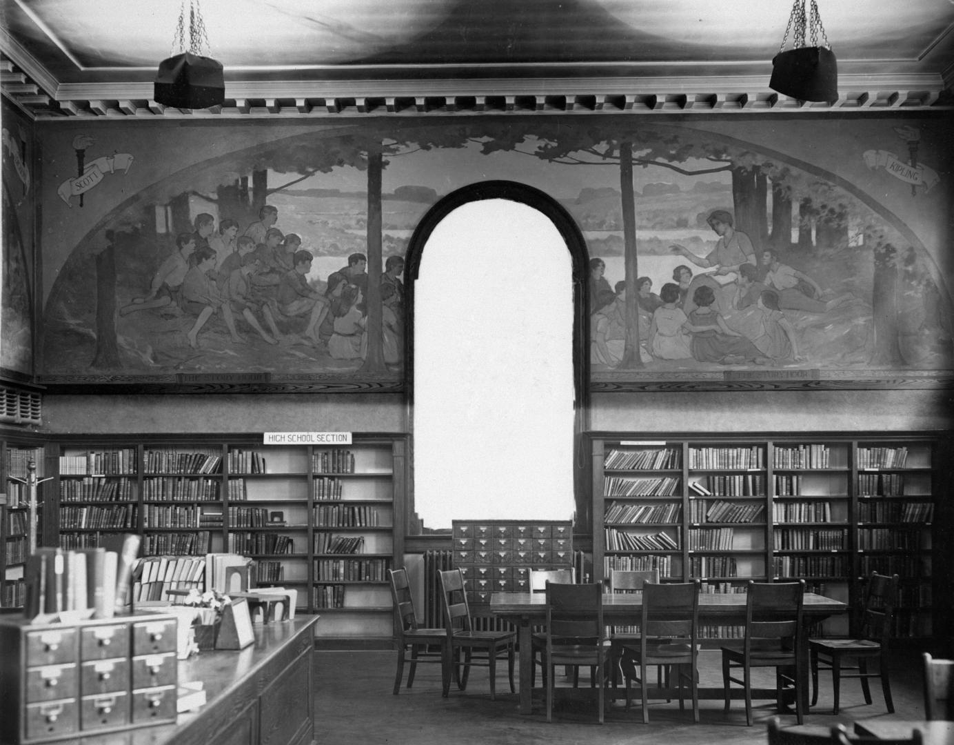 Image shows an interior of the library branch with book stacks along the walls, the staff desk  ...