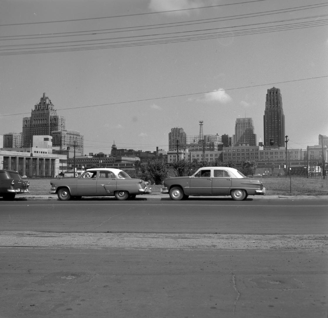 Image shows a street view with some cars on it and harbour buildings in the background.