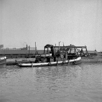 Image shows a tugboat on the water with a few buildings in the background.