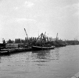 Image shows a docked tugboat with some cranes in the background.