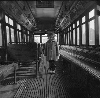  Image shows an interior of a rail car. There is a child standing in between the seats.
