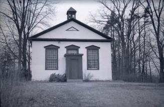 A photograph of a one story church, with a small bell tower or steeple and three windows above  ...