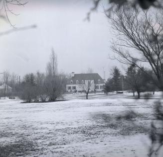 Image shows some land covered by snow. There is a house in the background.