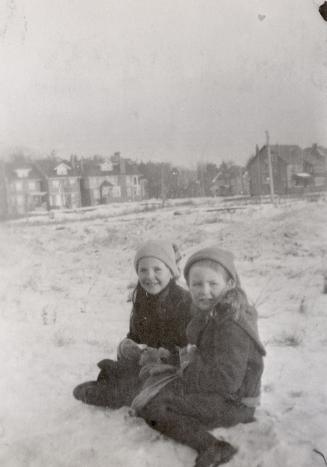 Image shows two girls sitting on the snow with some houses in the background.