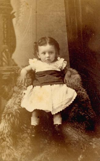 A black and white portrait of a girl of toddler age dressed in a white Victorian-era dress with ...