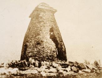 A photograph of a stone mill with a pointed tower, standing above a rocky area.