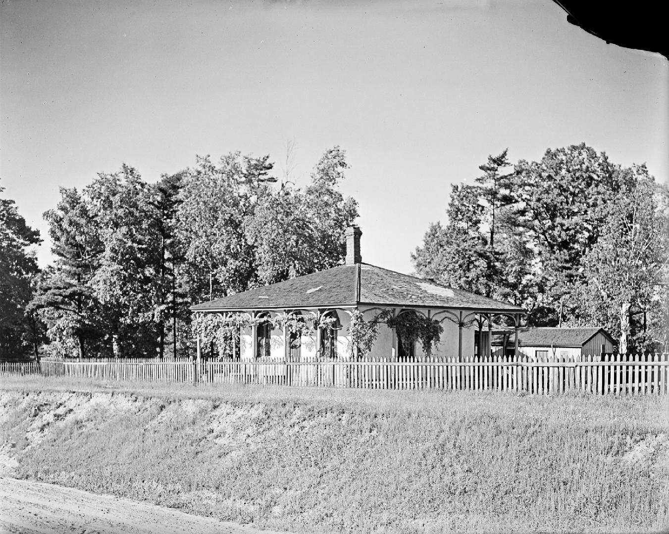 Image shows a cottage within a fenced area with some trees behind it.
