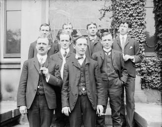 A formal group portrait of 8 men in three-piece suits and ties standing outdoors, on a small st ...
