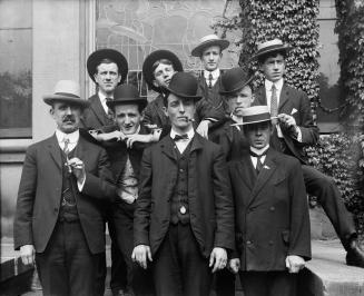An informal group pose: 9 men in three-piece suits, and hats, posed on a short outdoor staircas ...