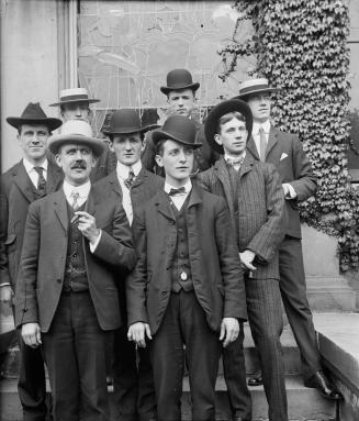 A formal, posed group portrait of 8 men, wearing hats and three-piece suits. They are posed on  ...
