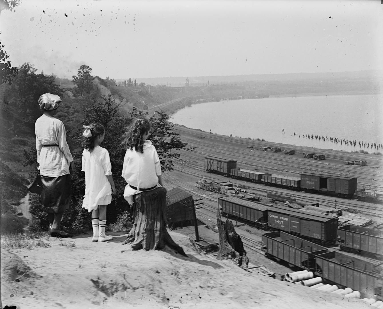 Three long-haired children in dresses, at the top of a ridge, gaze down at a railway yard on th ...