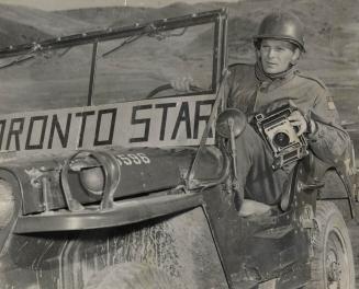 A man in army fatigues and a helmet sits, holding an old-fashioned camera, in an open jeep with ...
