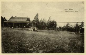 Photograph shows a small wooden building with a porch on a grassy hill.