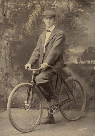 A young man in plus fours and a flat cap riding a safety bicycle.
