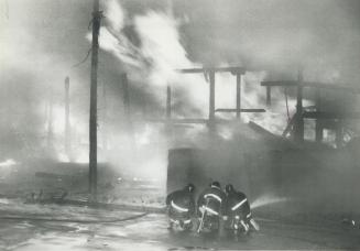 Image shows three firefighters working on scene while fire is destroying houses.