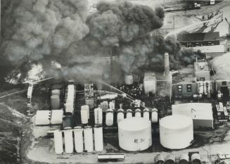 A column of smoke, visible over a large area of Metro, rises from the flaming ruins of the Ashland Oil Co