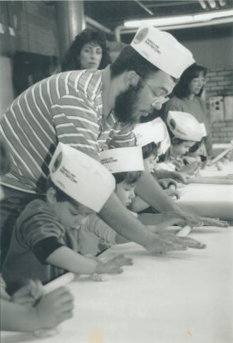 Black and white photo of man and children with paper hats rolling out pastry