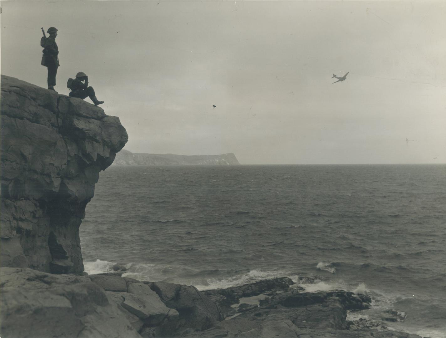 On a precipitous clift, to the right, members of the Canadian garrison watch a plane swoop in from a search for U-boats