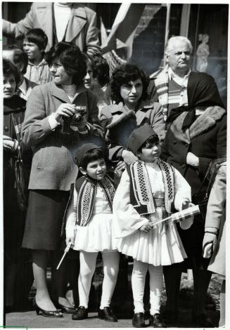 Two children dressed in white pleated skirts and tights, smocked vests and black caps, stand in ...