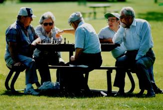 A group of men watch two chess players, all are sitting on a picnic table