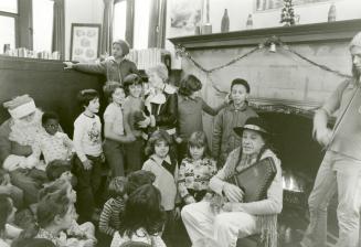 Image shows a singer and Santa Claus seated by the fireplace with a group of children around th ...