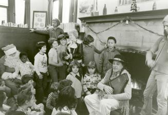 Image shows a singer and Santa Claus seated by the fireplace with a group of children around th…
