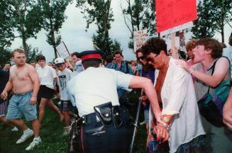 Bare-chested man watches as police lead away Adele Arnold, one of six women arrested yesterday for baring their breasts in a Waterloo park. Minutes la(...)
