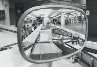 The view in the mirror at a postal encoder's desk shows letters waiting to be stamped with a bar-shaped code for machine sorting. The long desks of en(...)
