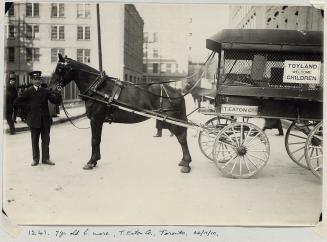 Eaton's delivery agent holding bridle of a horse harnessed to a wagon that says &quot;Toyland w ...