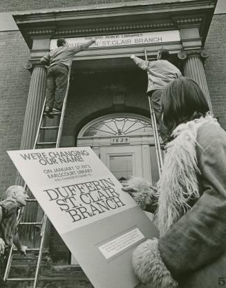Woman watches two men on ladders install a library sign above entrance reading 'Toronto Public  ...