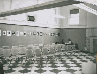 Three rows of empty chairs arranged on a checkerboard pattered floor. A row or artwork hangs on ...