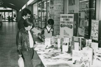 A man and two boys browse a book display at a table in the corridor of a shopping mall.