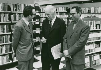 Three men in suits, one with papers in hand, congregate in front of a book shelf with a sign re ...