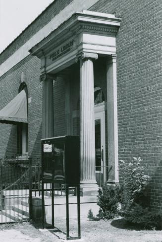 Side view of library entrance flanked by cement pillars . A black display case stands right of  ...