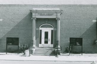 Front entrance of library with arched door flanked by cement pillars. Black display cases stand ...