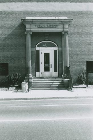 Front entrance of library. Steps lead to an arched door flanked by cement pillars. Sign above d ...
