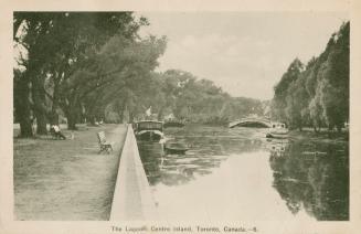 Black and white picture of a lagoon with boats and bridge and a park bench looking over the lag ...