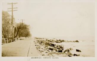 Black and white picture of wooden boardwalk with telephone poles and rocks and lake. 
