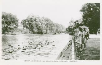 Black and white picture of a group of three girls standing on a dock watching ducks swimming in ...