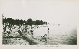 Photo of crowds of people on a beach with some wading or swimming in the water. 