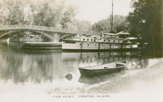 Photo of a fireboat docked at wharf with bridge in background and small row boat in foreground.…